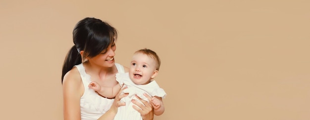 Happy cheerful young mother playing with cute baby on brown studio background